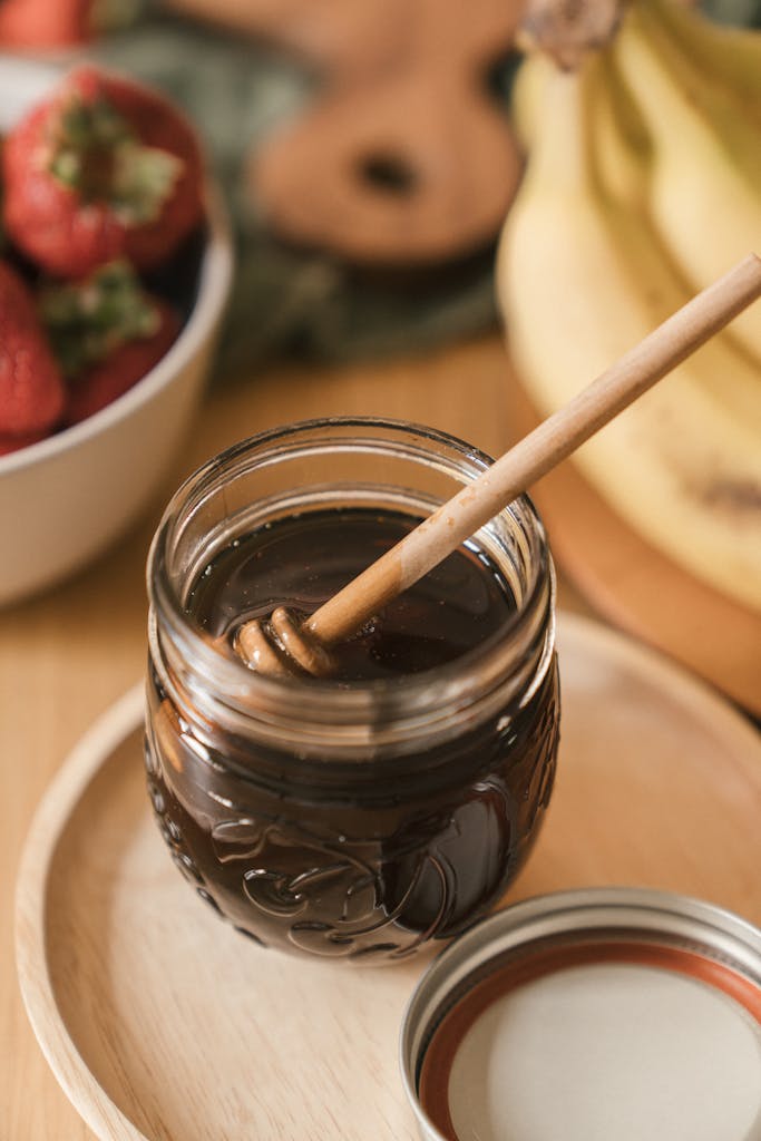 A glass jar of honey with a dipper on a wooden plate surrounded by fresh fruits.