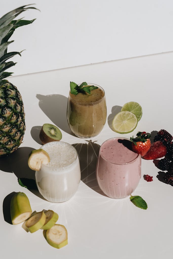 Three vibrant fruit smoothies surrounded by fresh tropical fruits on a sunlit surface.