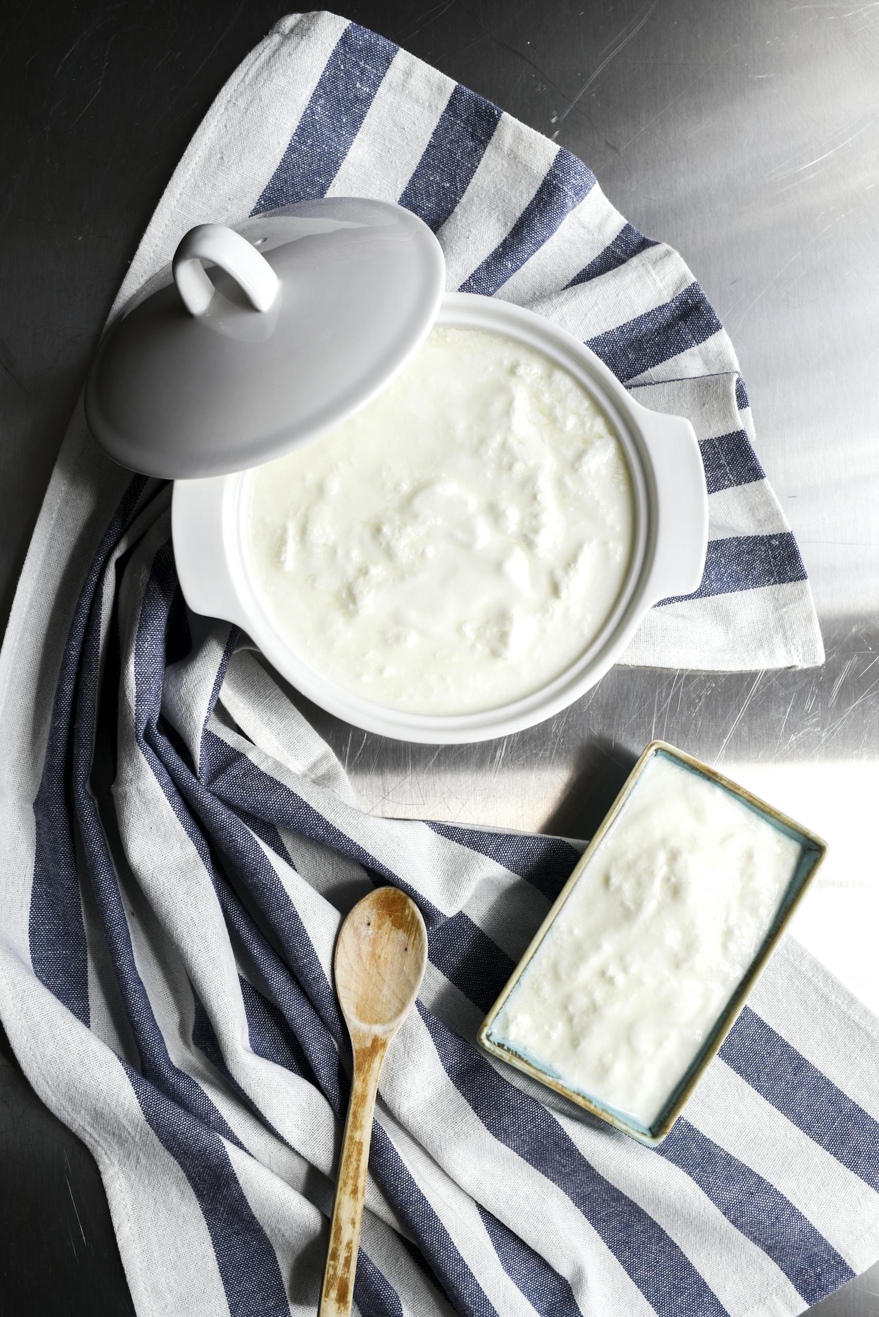 Top view of homemade yogurt in a ceramic pot, styled with a wooden spoon and striped cloth.